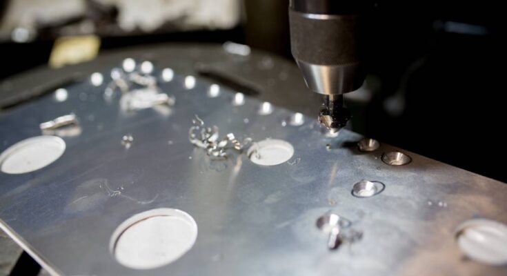 A machine drilling circular holes into a sheet of metal. The resulting metal burrs are off to the side.