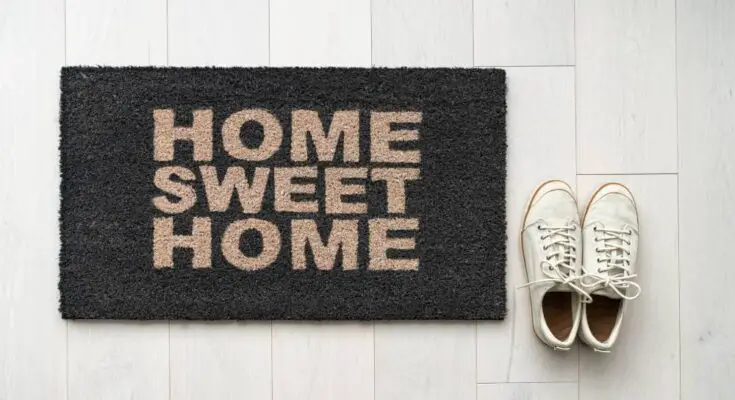 Black welcome rug with tan font that says “Home Sweet Home” on top of a white wood floor next to a pair of shoes