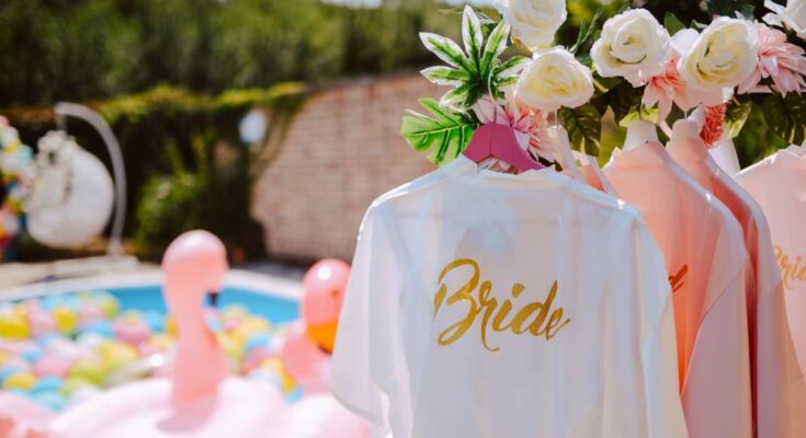Pink bridesmaid robes hanging behind a white robe labeled "bride", and a pool with flamingo floats in the background.