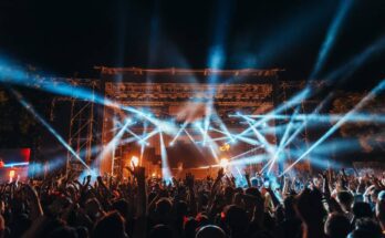 A large crowd of people with their hands in the air in front of a stage with tons of lights at a music festival.