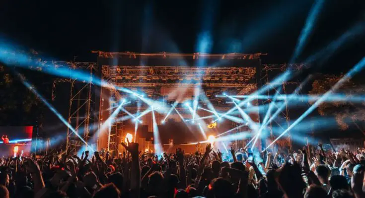 A large crowd of people with their hands in the air in front of a stage with tons of lights at a music festival.