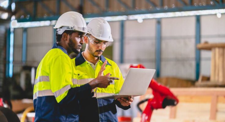 Two men standing in white hard hats and high-vis clothing working with a laptop on a manufacturing floor.