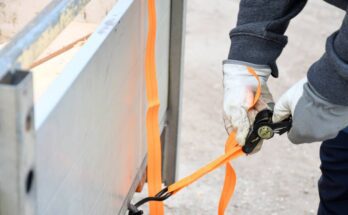 A logistics professional wearing work gloves holds a tensioning device to secure orange rachet straps to a truck trailer.