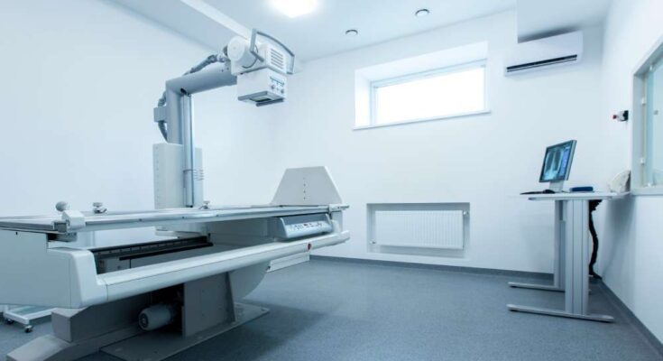 An immaculate and brightly lit radiology room in a hospital featuring an X-ray table and machine and computer monitor.