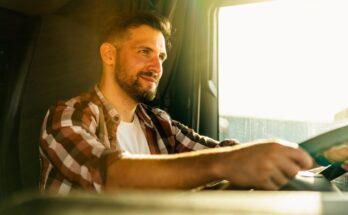 A truck driver wearing a white shirt and flannel, smiling with both hands on the steering wheel of his truck.