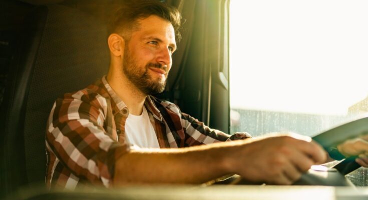 A truck driver wearing a white shirt and flannel, smiling with both hands on the steering wheel of his truck.