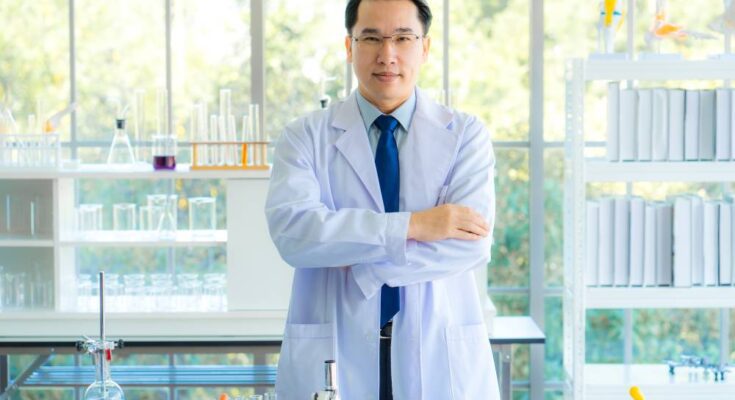 A lab manager stands in his laboratory with his arms folded. He wears a white coat, a blue tie, and glasses.
