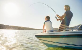 A father and son standing on a boat that is floating in a lake. The father is reeling in a catch with his fishing pole.