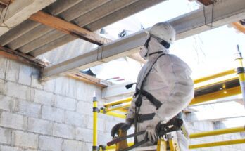 A worker in personal protective equipment stands on a yellow ladder inspecting an interior beam and wall.