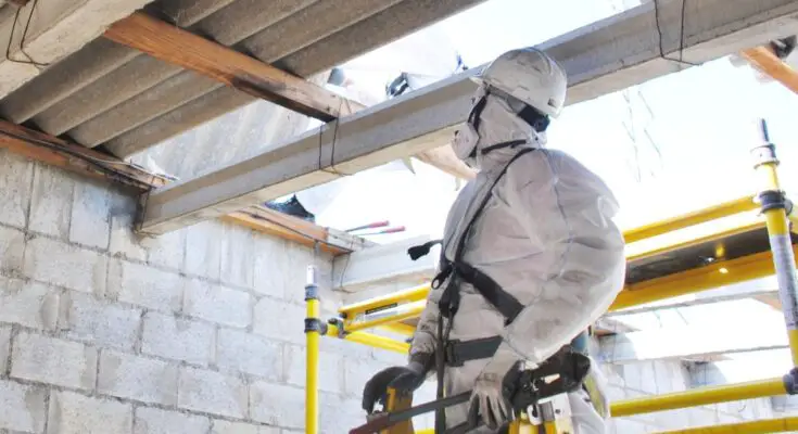 A worker in personal protective equipment stands on a yellow ladder inspecting an interior beam and wall.