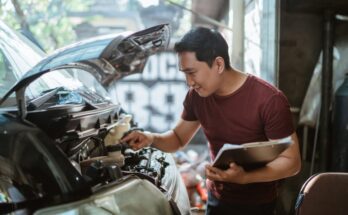 A young man wearing a burgundy shirt and holding a clipboard checks a silver vehicle's brake fluid at a car repair shop.