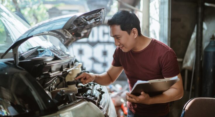 A young man wearing a burgundy shirt and holding a clipboard checks a silver vehicle's brake fluid at a car repair shop.