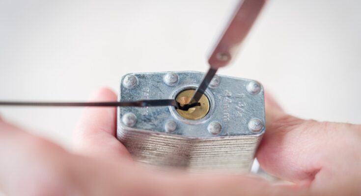 The bottom of a silver lock with a gold keyhole getting picked with a tension wrench and a pick.