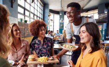 A young male server is serving a table of women their meals. The women are looking at the server and smiling.