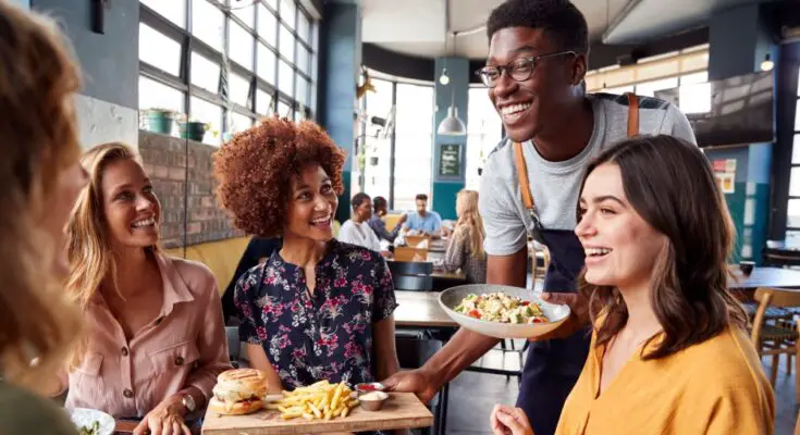 A young male server is serving a table of women their meals. The women are looking at the server and smiling.