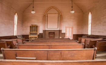 An old church with rows of rustic wooden pews, white wood-panel walls, and a small organ to the left of the pulpit.