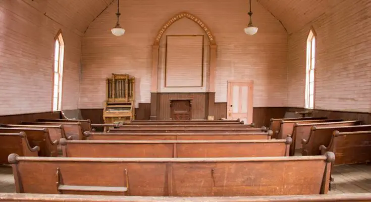 An old church with rows of rustic wooden pews, white wood-panel walls, and a small organ to the left of the pulpit.