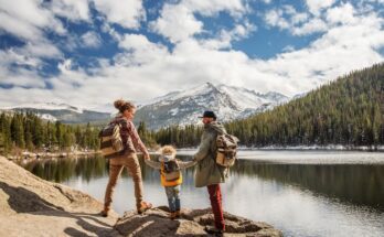 A mother and father holding their son's hands as they stand looking at a view of a lake and mountain on a hike.