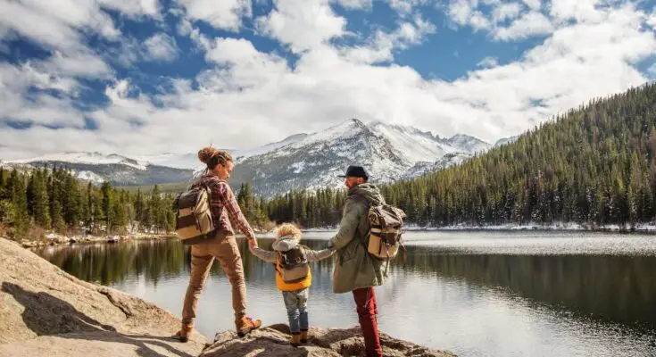 A mother and father holding their son's hands as they stand looking at a view of a lake and mountain on a hike.