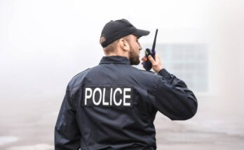 A bearded man wearing a black baseball cap and jacket that reads “police” speaks into a handheld radio.