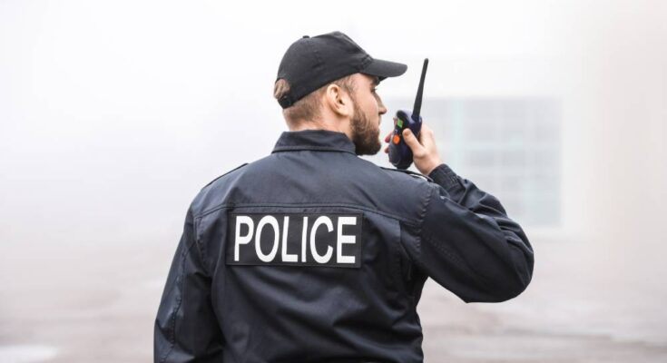 A bearded man wearing a black baseball cap and jacket that reads “police” speaks into a handheld radio.