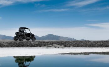 A black utility task vehicle cruising along the water, with a stunning mountain range in the background.