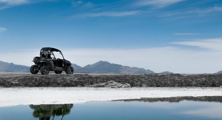 A black utility task vehicle cruising along the water, with a stunning mountain range in the background.