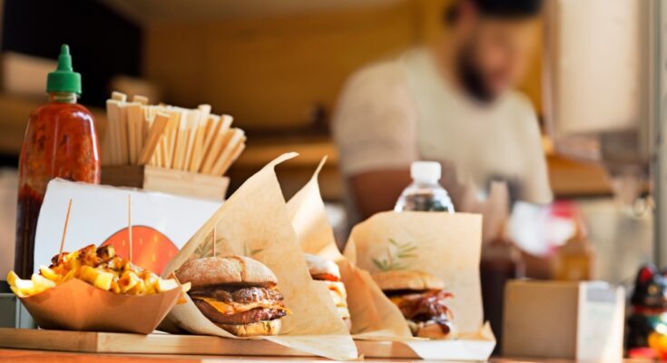 A chef cooks in a food truck while three full meals sit at the window, waiting for the guests to pick them up.