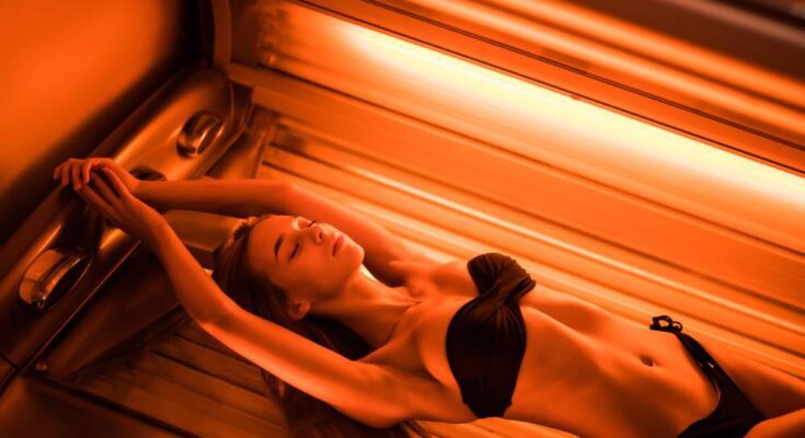 A young woman wearing a bikini, lying with her arms above her head in a tanning bed with orange lights on.