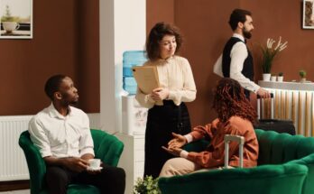 A hotel manager talks to two hotel guests sitting in green velvet chairs. One has a suitcase and the other's drinking coffee.