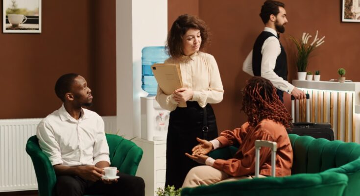 A hotel manager talks to two hotel guests sitting in green velvet chairs. One has a suitcase and the other's drinking coffee.