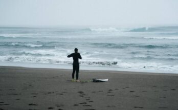 A man wearing a black wet suit, standing on the beach with a surfboard leash attached to his ankle and facing a stormy ocean.