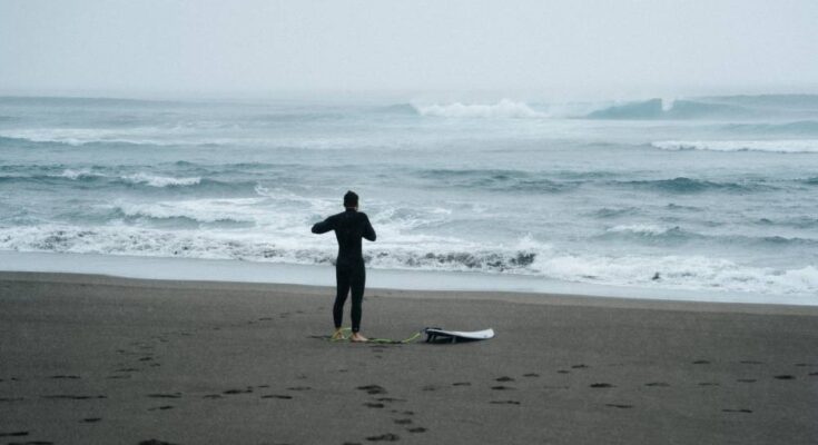 A man wearing a black wet suit, standing on the beach with a surfboard leash attached to his ankle and facing a stormy ocean.