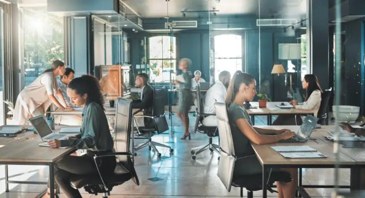 A busy office with multiple desks and chairs occupied by well-dressed employees working on their laptops.