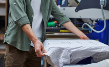 An unidentifiable male is placing a white t-shirt onto a screen printing machine in his small business workspace.