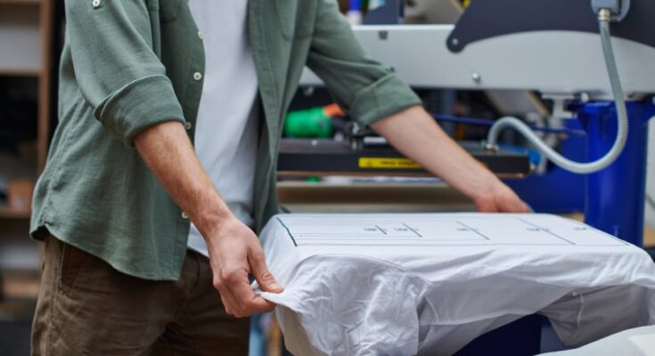 An unidentifiable male is placing a white t-shirt onto a screen printing machine in his small business workspace.