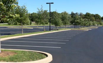 An empty asphalt parking lot on a sunny day. There are lush trees and tall light posts throughout the parking area.