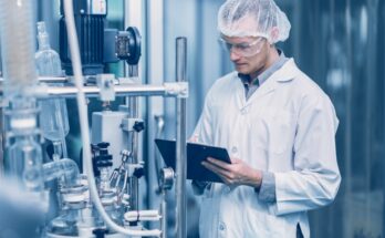 A male employee wears a white lab coat, goggles, and a hair net while writing notes on a clipboard.