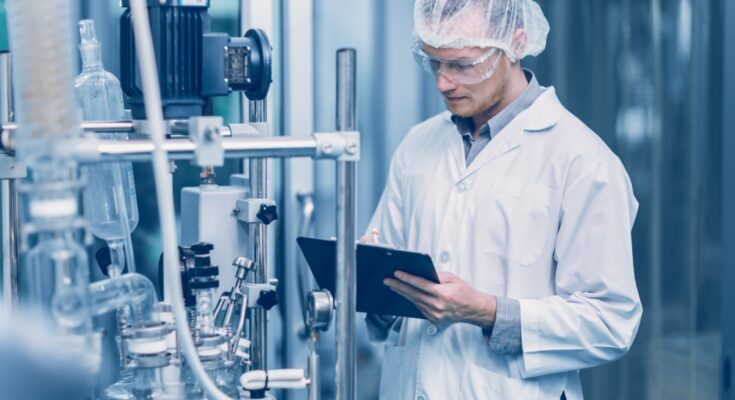 A male employee wears a white lab coat, goggles, and a hair net while writing notes on a clipboard.