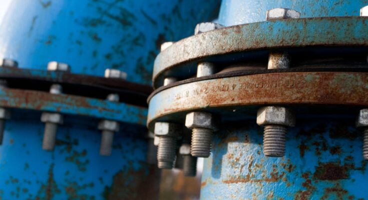 A pair of blue pipelines run next to each other, featuring rust on the outside. Nuts and bolts hold them together.