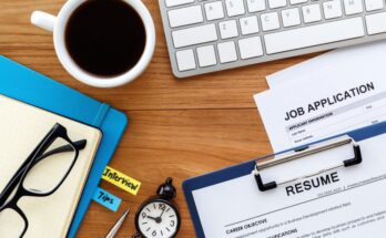 A wooden desk with a laptop, cup of coffee, pair of glasses, timer, binder of interview tips, resume, and job application on it.