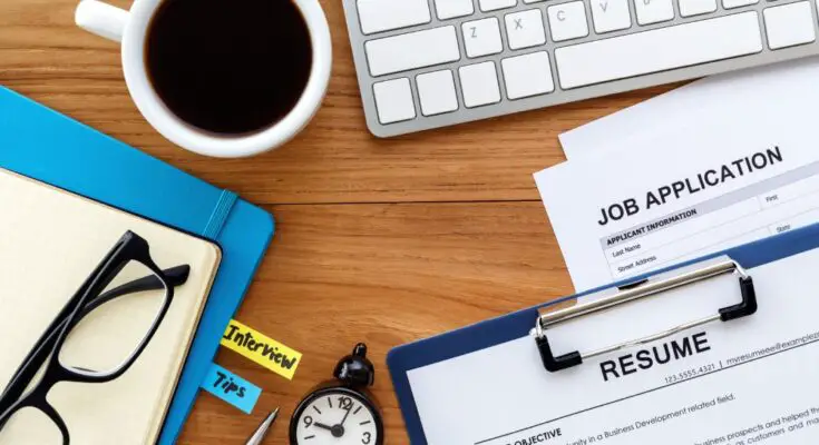 A wooden desk with a laptop, cup of coffee, pair of glasses, timer, binder of interview tips, resume, and job application on it.