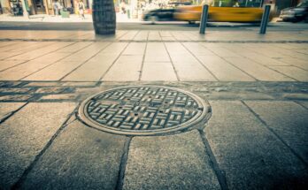 An intricately designed manhole cover on a sidewalk in a bustling city. There's a taxi driving by and pedestrians nearby.