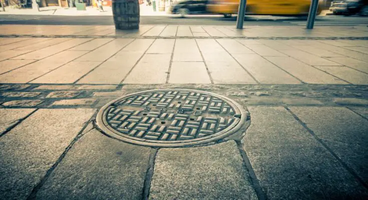 An intricately designed manhole cover on a sidewalk in a bustling city. There's a taxi driving by and pedestrians nearby.