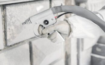 A white-gloved hand is up close and using a sandblaster tool to remove debris from a white brick wall.