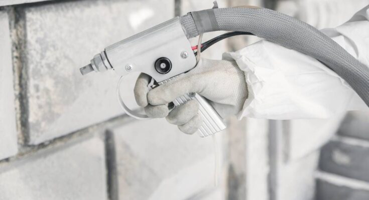 A white-gloved hand is up close and using a sandblaster tool to remove debris from a white brick wall.