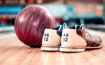 A red bowling ball and a pair of size 12 bowling shoes sitting on the ground in front of a bowing lane.