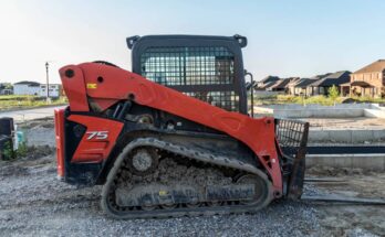 A red and black skid steer sits on a home building construction site. Built homes appear in the background.