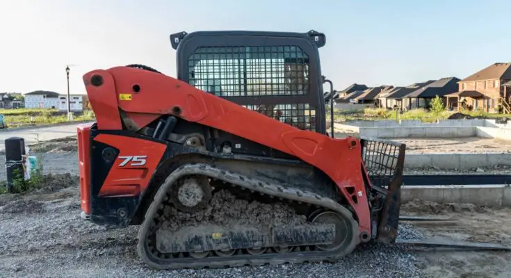 A red and black skid steer sits on a home building construction site. Built homes appear in the background.