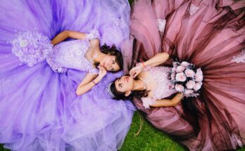 Two teenage girls in quinceañera dresses, one lilac and one dusty rose, lay in the grass and look at once another.